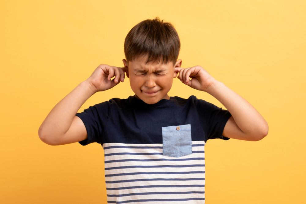 Portrait of angry unhappy irritated preteen boy wearing casual t-shirt covering plugging ears with fingers and squinting isolated on yellow studio background, scared of loud noise voice, bad behaving